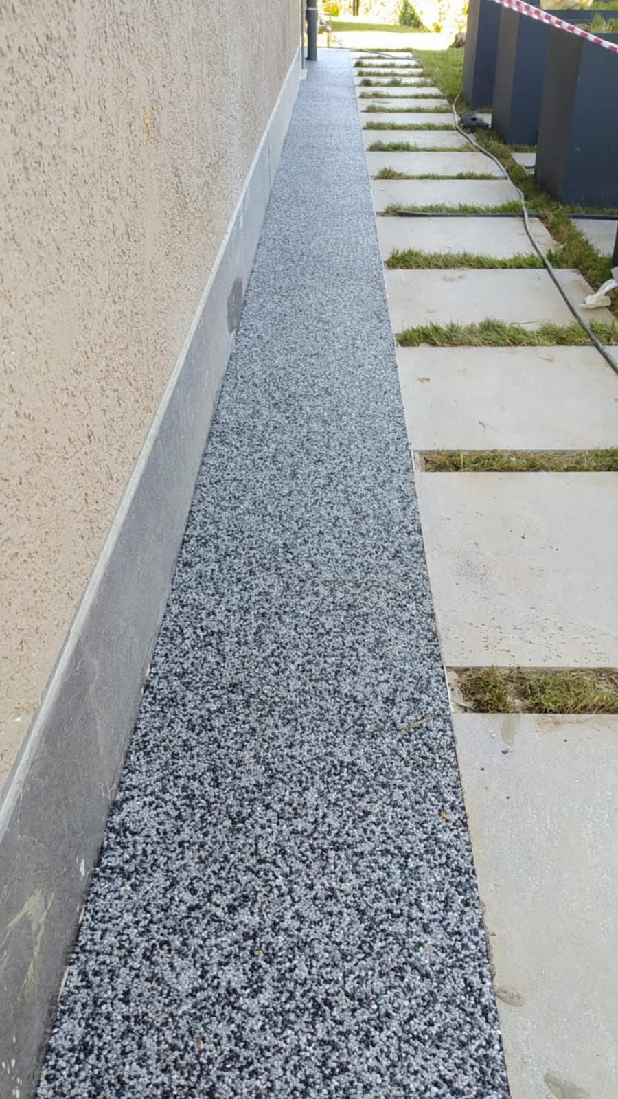 Bina duvarı boyunca uzanan dar yürüyüş yolunda gri taş halı; yanında çim aralıklı beton plakalar